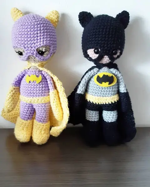 Dos amigurumis de superhéroes con capa y antifaz, uno amarillo y morado y otro gris y negro