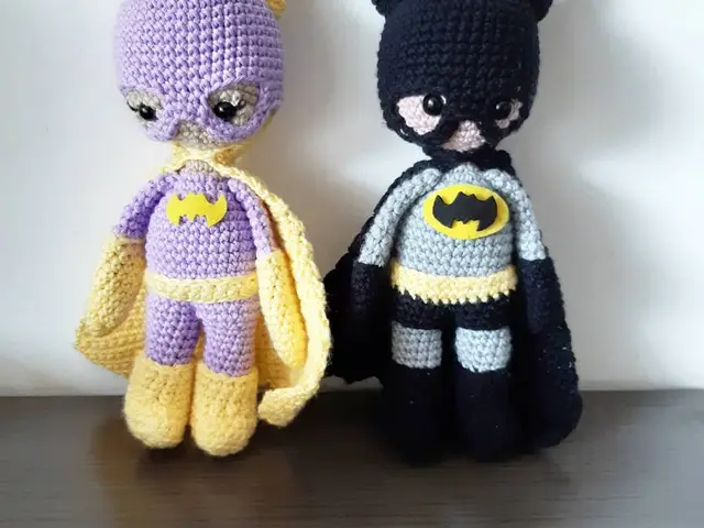 Dos amigurumis de superhéroes con capa y antifaz, uno amarillo y morado y otro gris y negro