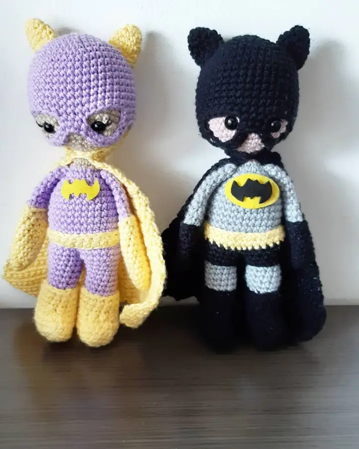 Dos amigurumis de superhéroes con capa y antifaz, uno amarillo y morado y otro gris y negro