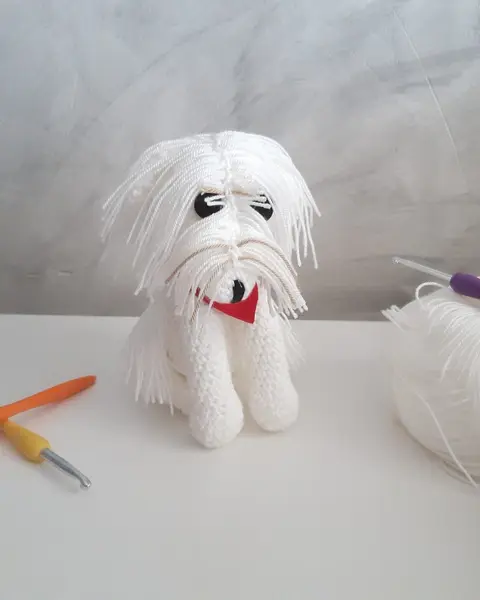 Amigurumi de bichón maltés blanco con lazo azul, tejido a crochet, junto a un ovillo y una aguja