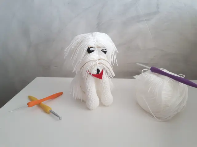 Amigurumi de bichón maltés blanco con lazo azul, tejido a crochet, junto a un ovillo y una aguja