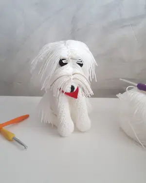 Amigurumi de bichón maltés blanco con lazo azul, tejido a crochet, junto a un ovillo y una aguja