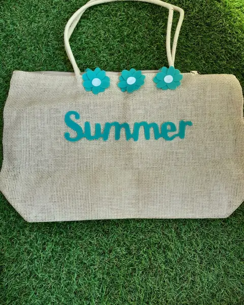 Bolso de paja beige con la palabra Summer y flores de fieltro turquesa cosidas, sobre césped