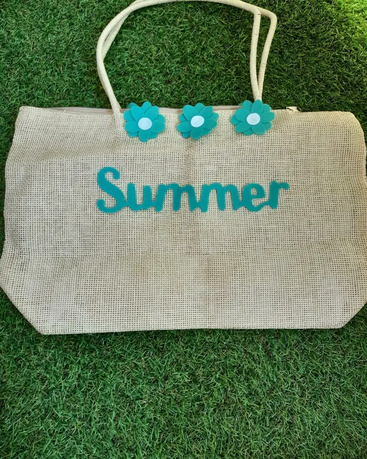 Bolso de paja beige con la palabra Summer y flores de fieltro turquesa cosidas, sobre césped