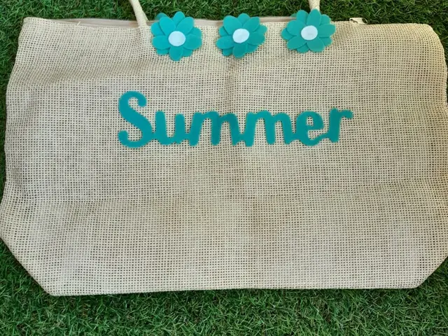 Bolso de paja beige con la palabra Summer y flores de fieltro turquesa cosidas, sobre césped