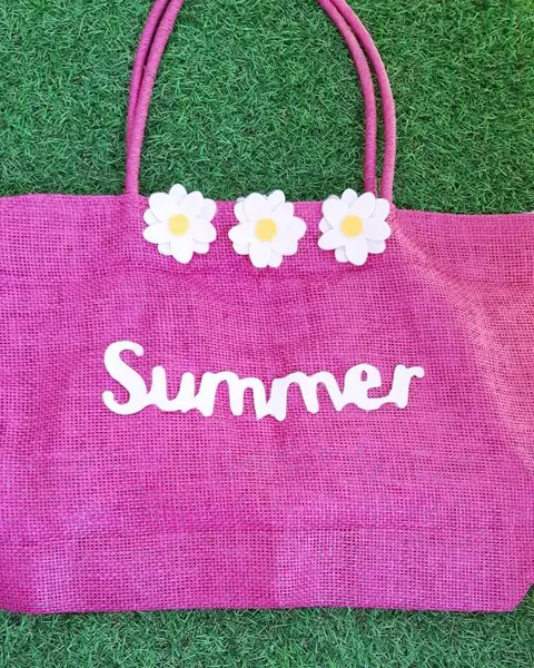 Bolso de paja fucsia con la palabra Summer y flores de fieltro blanco cosidas, sobre césped