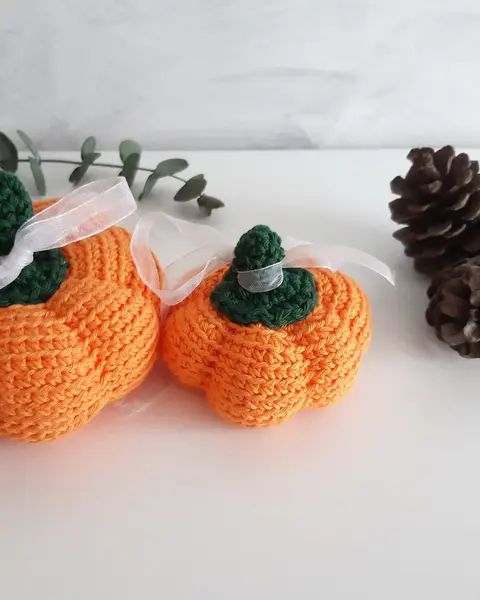 Tres calabazas de crochet naranjas de distinto tamaño con rabito verde, junto a piñas