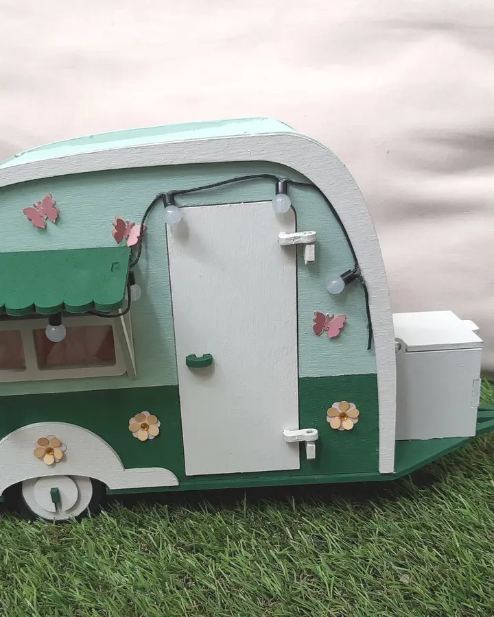 Maqueta de caravana de madera pintada en verde y crema, con toldo y ventanas, sobre césped