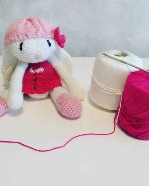 Amigurumi de una conejita con vestido rosa y lazo, tejida a crochet, sentada sobre fondo claro