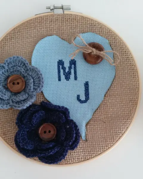 Bastidor de madera con iniciales bordadas en punto de cruz dentro de un corazón y flores de crochet