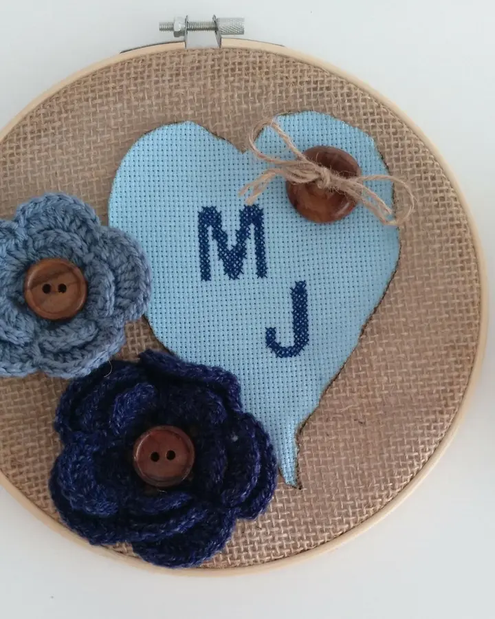 Bastidor de madera con iniciales bordadas en punto de cruz dentro de un corazón y flores de crochet