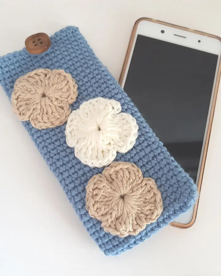 Funda de móvil de crochet azul cielo con flores tejidas, junto a un teléfono