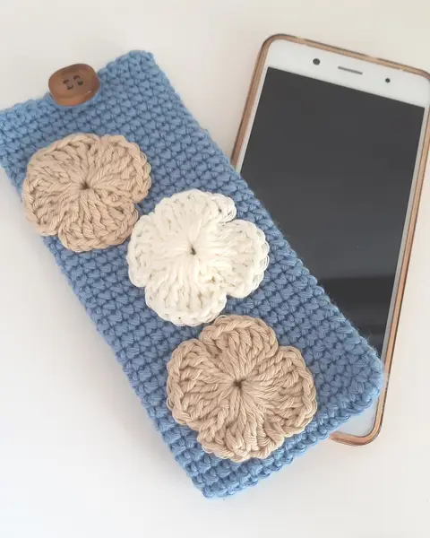 Funda de móvil de crochet azul cielo con flores tejidas, junto a un teléfono