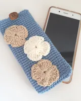Funda de móvil de crochet azul cielo con flores tejidas, junto a un teléfono