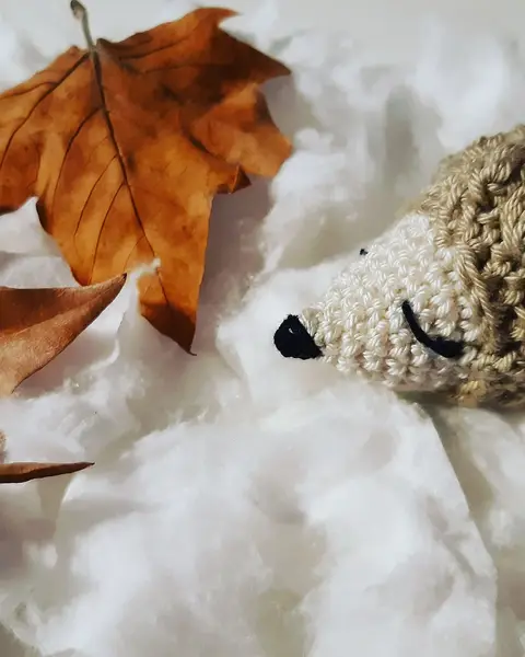 Llavero de crochet con forma de erizo beige y marrón con ojos cerrados junto a hojas secas de otoño