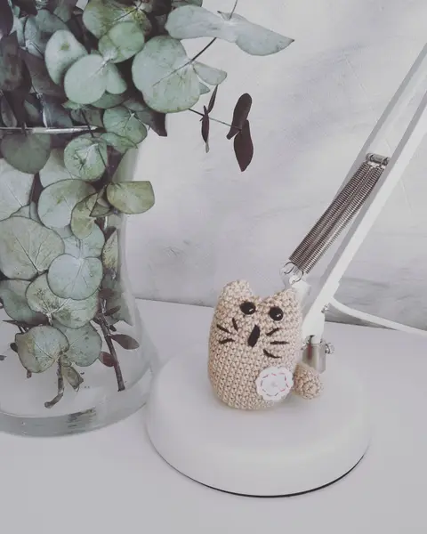 Llavero de crochet con forma de gato pequeño en hilo crudo, sentado sobre un escritorio blanco