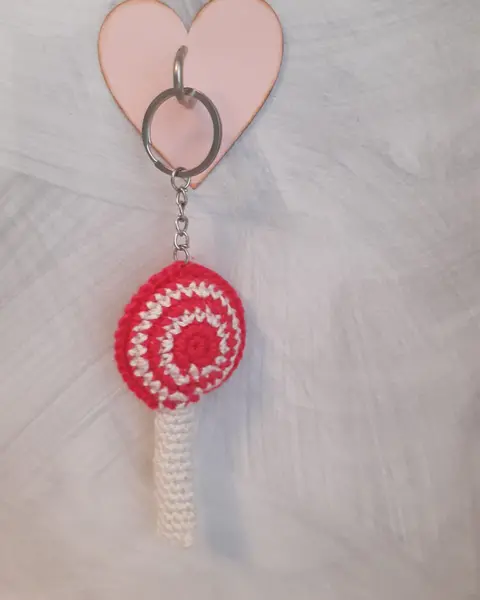 Llavero de crochet con forma de piruleta en espiral roja y blanca colgado de una anilla con corazón rosa