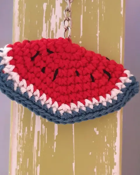 Llavero de crochet con forma de rodaja de sandía roja con pepitas negras y corteza verde, sobre madera pintada