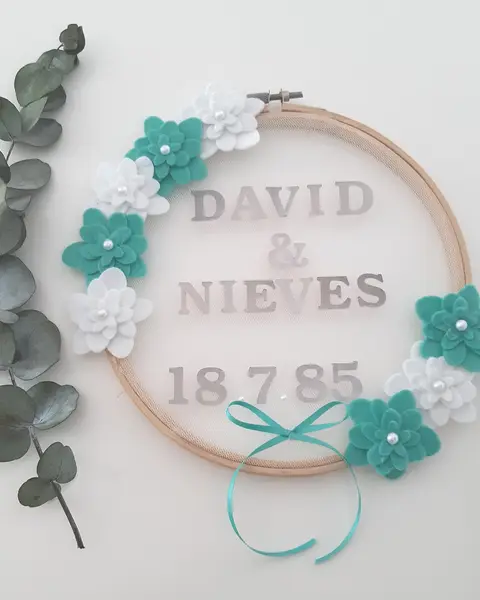 Porta alianzas en bastidor con tul blanco, flores de fieltro turquesa, eucalipto y nombres de los novios