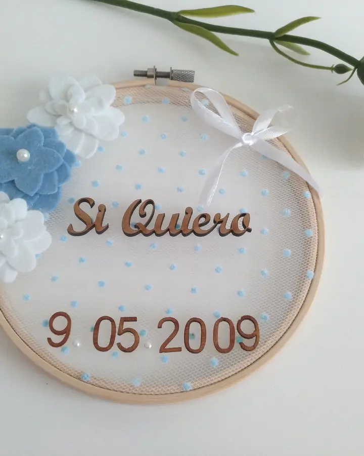 Porta alianzas en bastidor con tul azul, flores de fieltro azules y blancas y texto Sí Quiero en madera