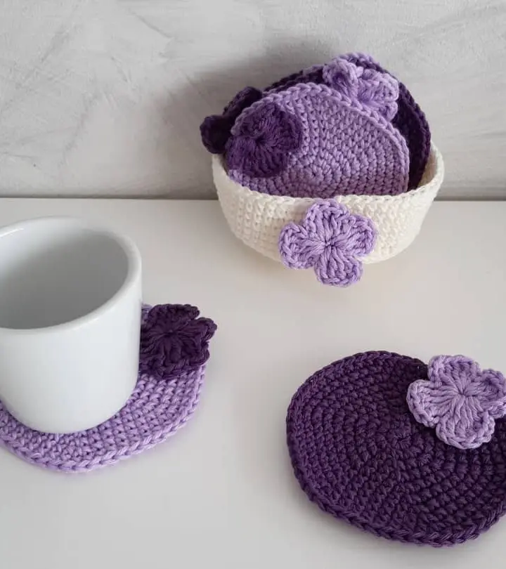 Posavasos redondos de crochet lila y morado con flores, junto a su cestita y una taza blanca