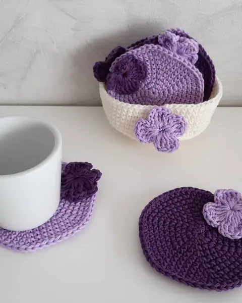 Posavasos redondos de crochet lila y morado con flores, junto a su cestita y una taza blanca