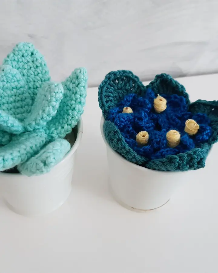 Dos suculentas de crochet, verde agua y azul con flores amarillas, en macetas de cerámica blanca