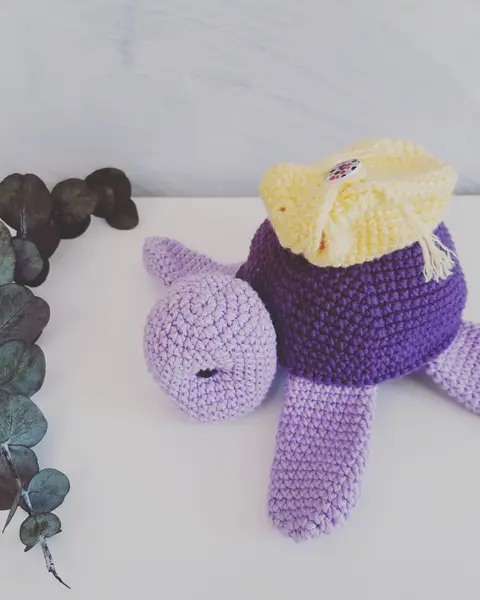 Tortuga amigurumi lila con caparazón morado y amarillo, con ojos de seguridad, junto a ramas de eucalipto
